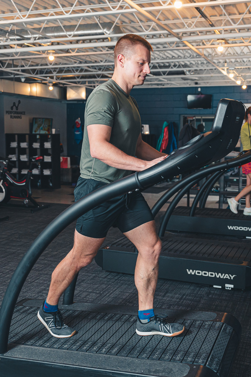 Person wearing a green t-shirt and black shorts walking on a treadmill at a performance running gym to warm up for his gait analysis to optimize your run performance