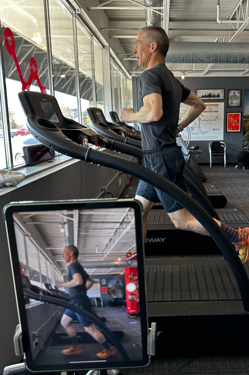 Person wearing a dark t-shirt and shorts working on optimizing their run performance with a gait analysis at Performance Running Gym. In the foreground is a phone taking a video of their running form
